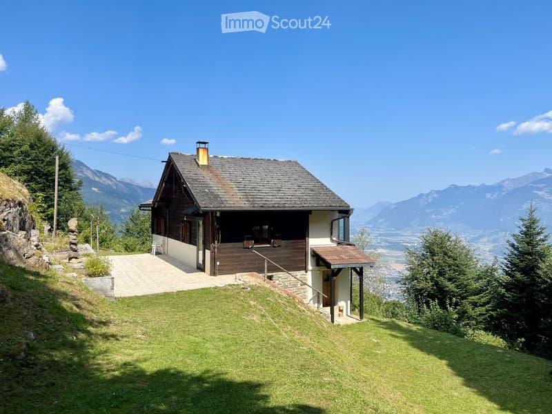 A two-story wooden house with a dark roof and white walls, situated on a grassy slope with a panoramic mountain view in the background.