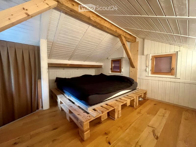 Cozy attic bedroom with wooden floors, slanted wooden ceiling, and a lofted bed on wooden pallets. Features a bench beneath the bed and a small window with blinds.
