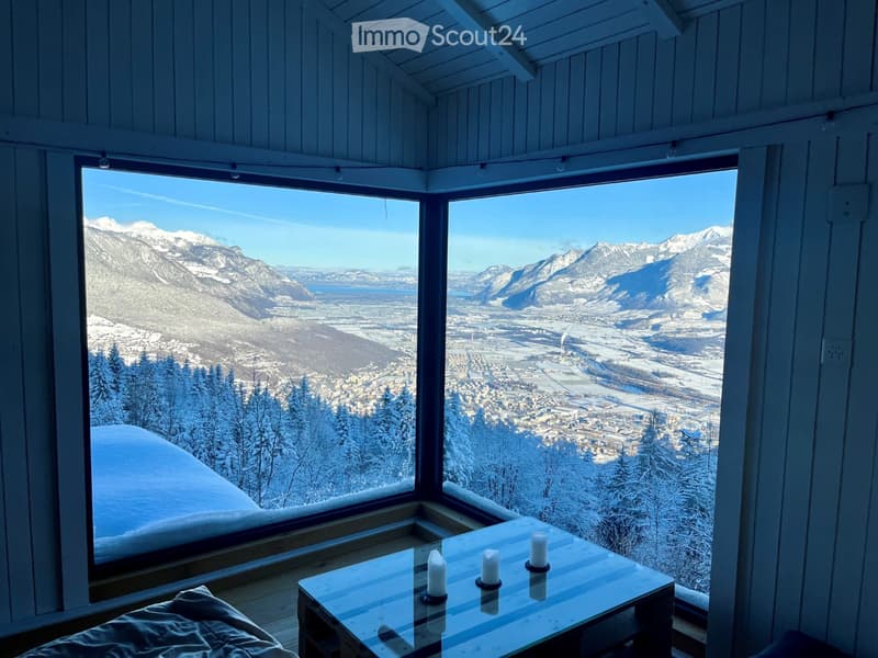 A spacious balcony with large windows offering a panoramic view of snow-covered mountains and a town below. The balcony features a glass table with candles and a cushioned seating area.