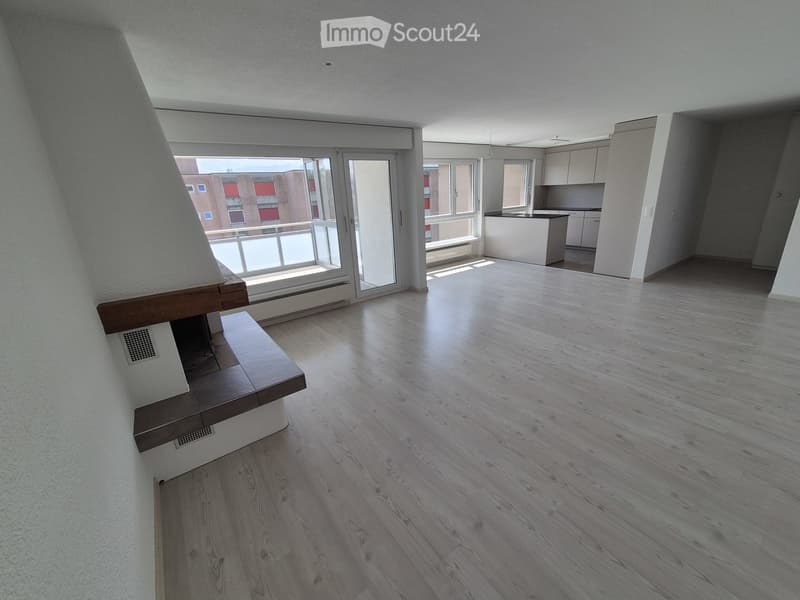 Empty room with light wooden flooring, white walls, and a large window leading to a balcony. The adjacent kitchen area is also visible with white cabinets and a countertop.