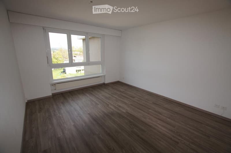 Empty room with wooden flooring, white walls, and a large window offering a view of the outside area.