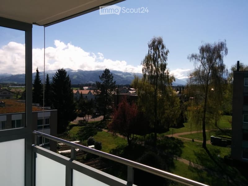 Balcony with a scenic view of a park, trees, and distant mountains under a clear blue sky.