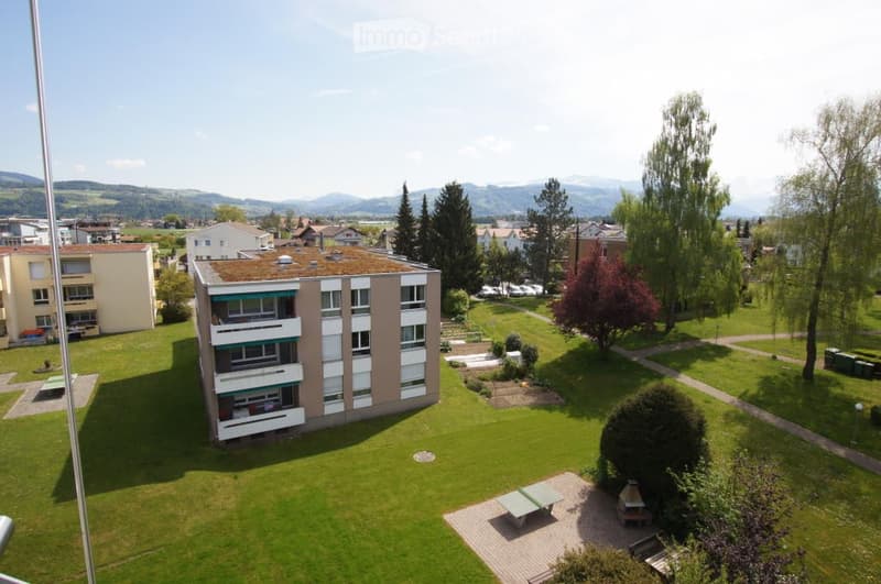 Multi-story residential building with balconies and a green lawn area in front, surrounded by trees and a scenic mountain backdrop.