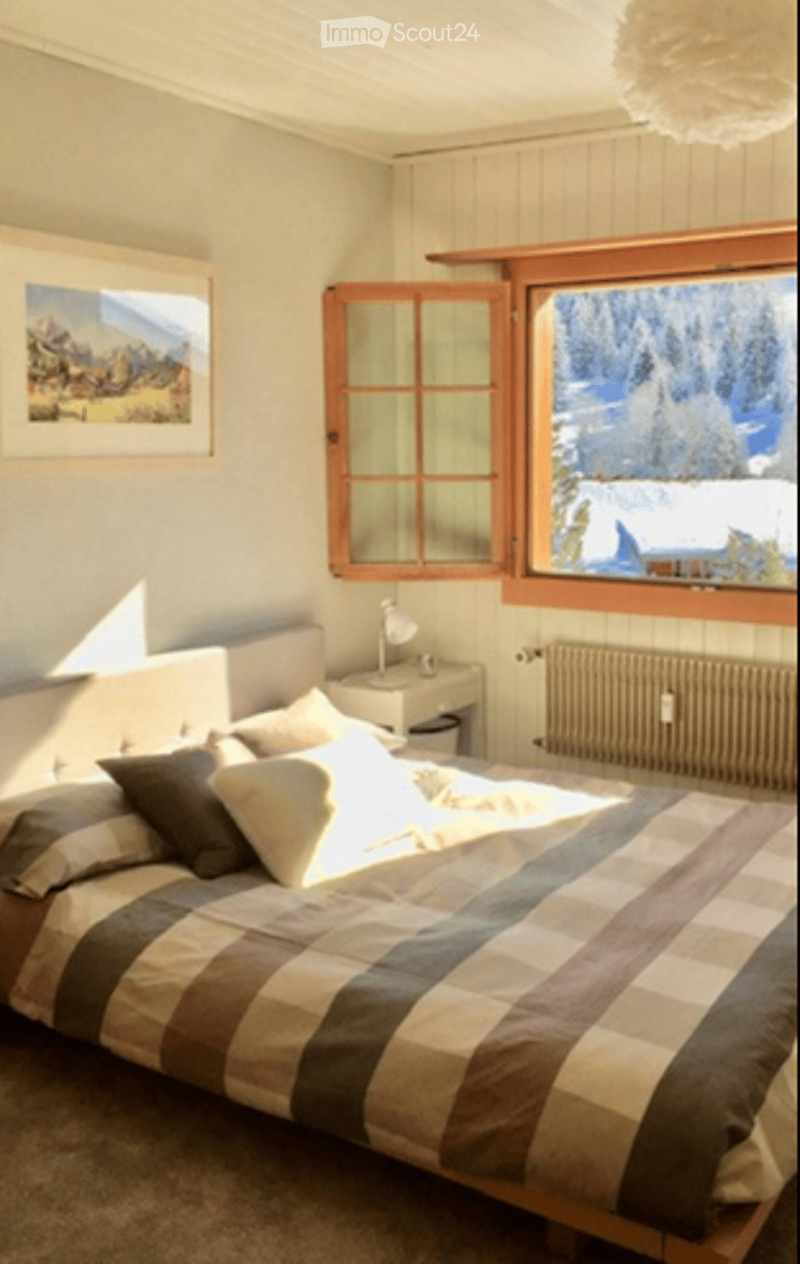 Cozy bedroom with a large window offering a scenic view, a neatly made bed with pillows and a blanket, a nightstand with a lamp, and a picture frame on the wall.