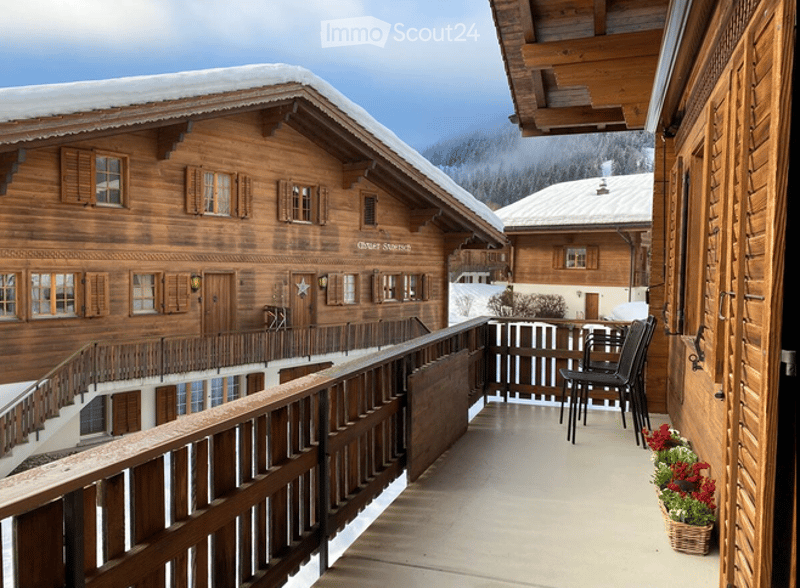 Balcony with wooden railings, overlooking a snow-covered wooden chalet with multiple windows and a staircase leading up to it.