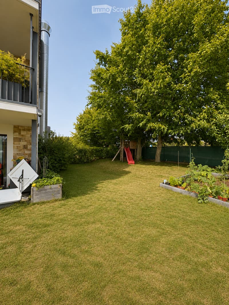 Well-maintained garden with a grassy lawn, a large tree, a red slide, and a raised garden bed with plants.