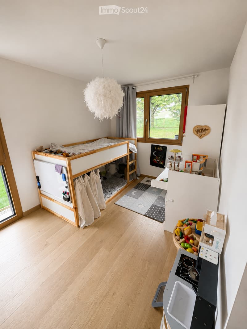 A well-lit room with a bunk bed, a small kitchen area, and a toy shelf. The room features wooden flooring, large windows, and a decorative hanging lamp.