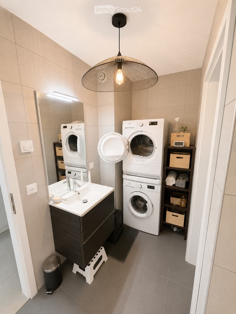 Modern bathroom with a sink, mirror, washing machine, dryer, and shelves holding towels and cleaning supplies.
