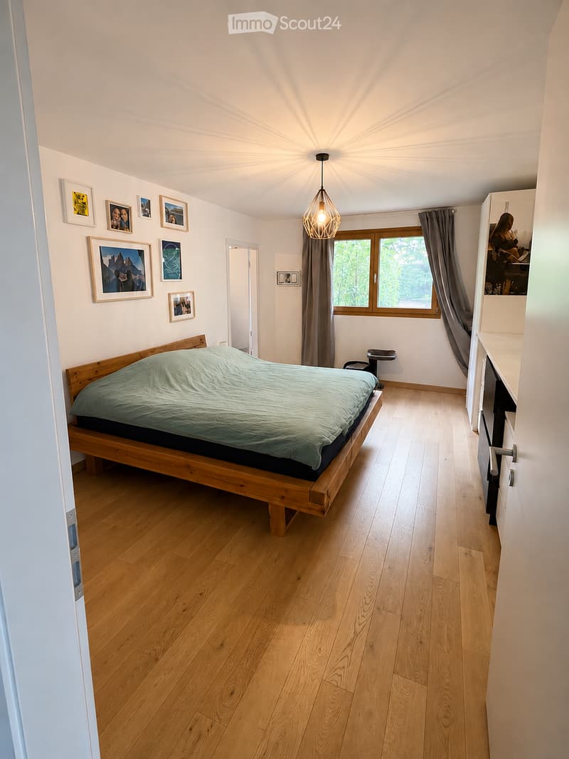 Bright bedroom with wooden flooring, a large bed with green bedding, a wardrobe, picture frames on the wall, and a pendant light fixture.