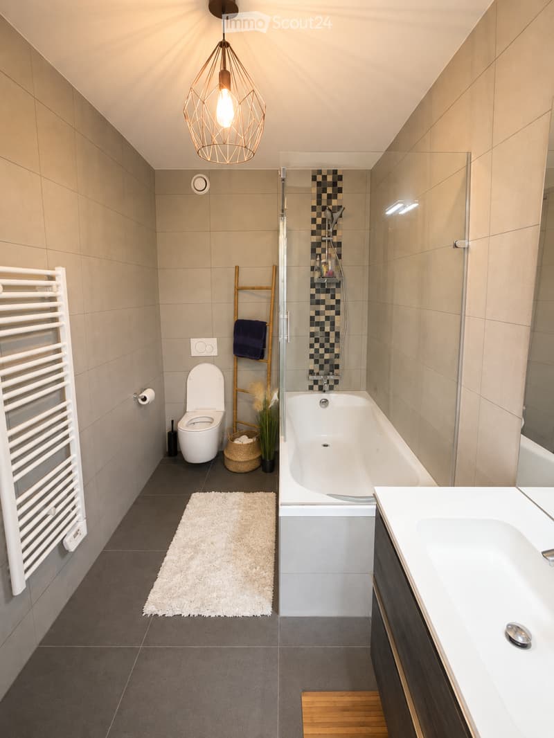 Modern bathroom with a bathtub, toilet, sink, radiator, and a shower area with a glass enclosure. Includes a white towel, a small rug, a plant, and a woven basket.