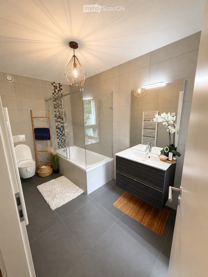Modern bathroom with a bathtub, toilet, sink, and mirror. Features include a wicker basket, wooden shelf, and a white rug on the floor.