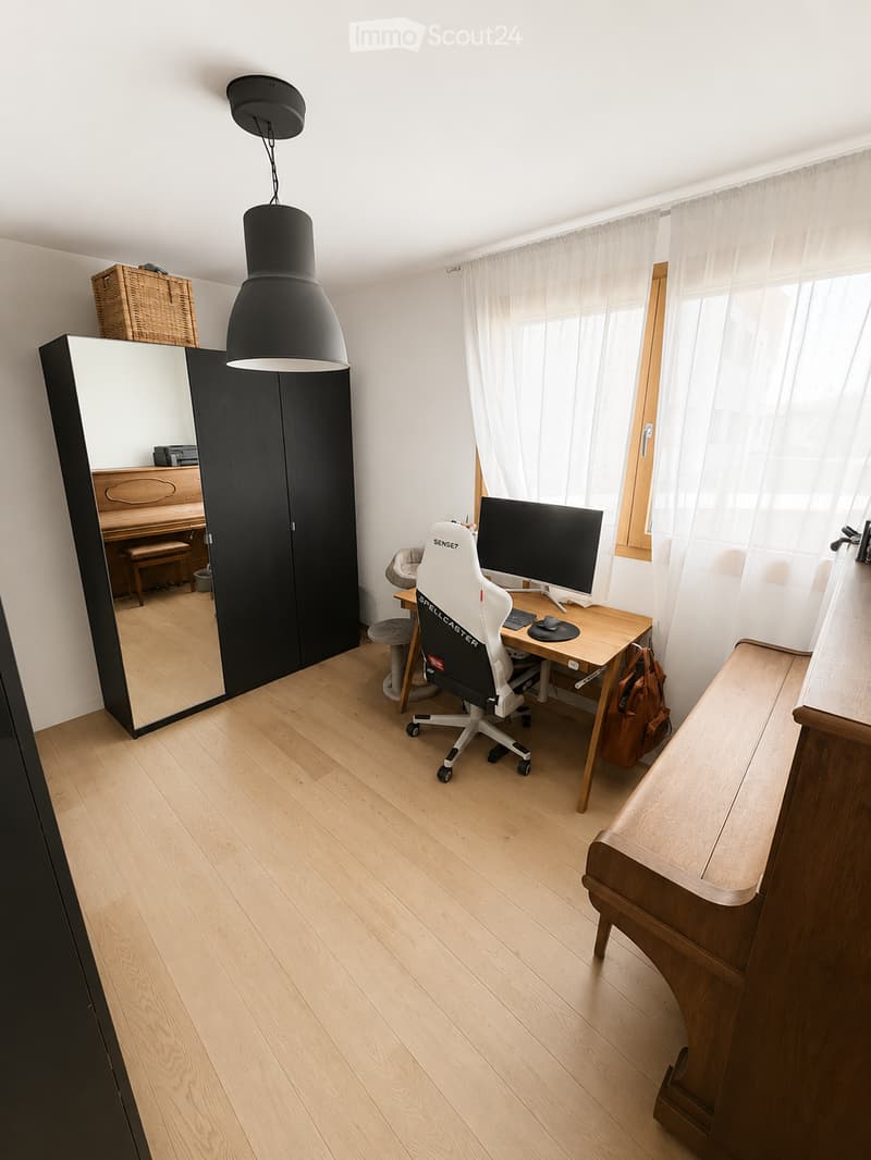 A well-lit room with wooden flooring, a large mirror, a desk with a computer and chair, a piano, and a wicker basket on top of the mirror.