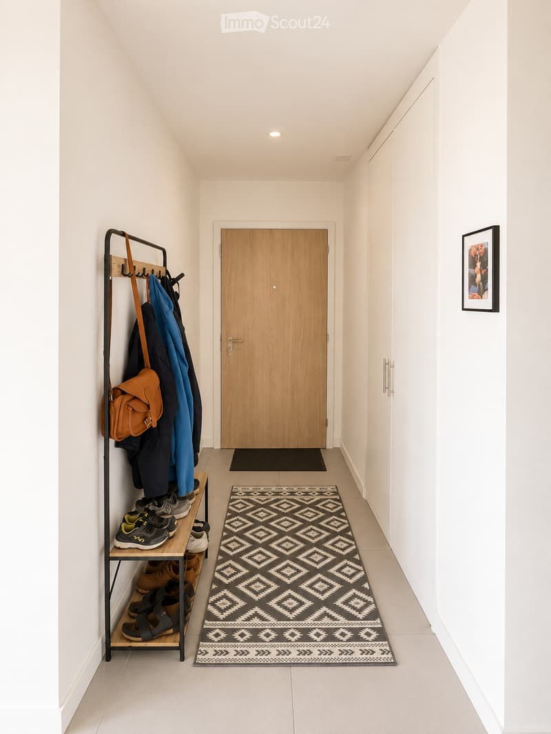 Modern hallway with white walls, tiled floor, and a patterned rug. Features a coat rack with jackets, shoes, and a handbag, along with a framed picture on the wall.