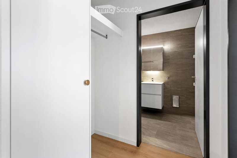 A modern hallway with white walls and wooden flooring, leading to a contemporary bathroom featuring a white vanity, wooden wall panels, and a sleek mirror.