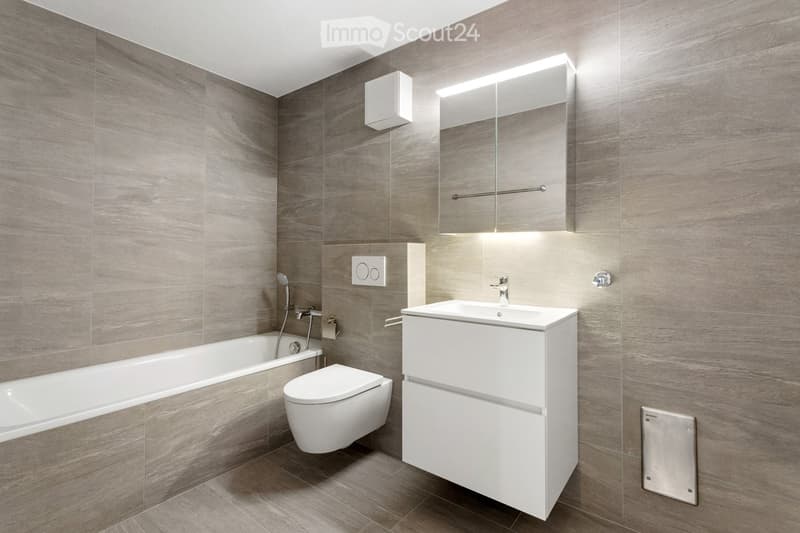 Modern bathroom with grey tiled walls and floor, featuring a bathtub, toilet, sink with a modern cabinet, and a large mirror with built-in lighting.