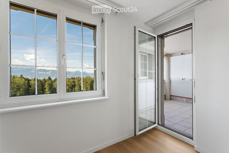 Empty room with white walls, wooden flooring, large windows, and an open sliding glass door leading to a balcony or terrace.