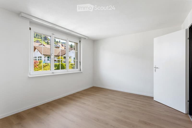 Empty room with white walls, wooden flooring, and a large window showing a view of houses and greenery outside.