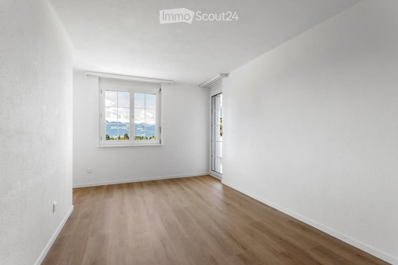 Empty room with white walls, wooden floor, large window with scenic view, and glass door leading outside.