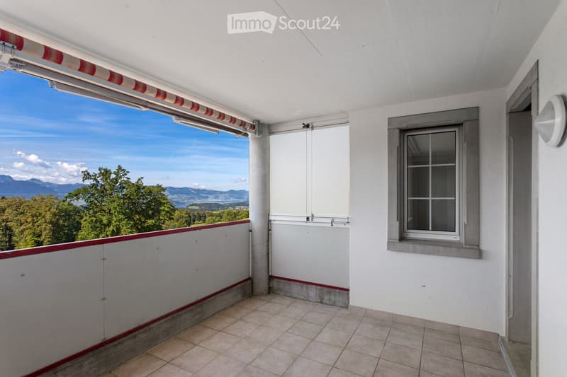 Balcony with tiled floor, white walls, large window with red and white striped awning, scenic mountain view.