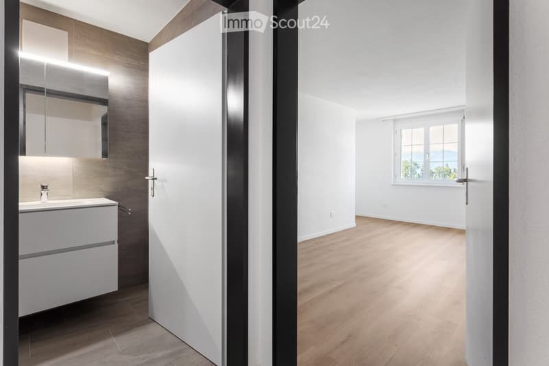 Empty room with light wooden flooring, white walls, and a large window with a scenic view. A modern bathroom is visible through an open doorway, featuring a sink, mirror, and sleek cabinetry.