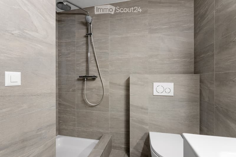Modern bathroom with grey tiled walls, a bathtub, toilet, and a handheld showerhead with a control valve.