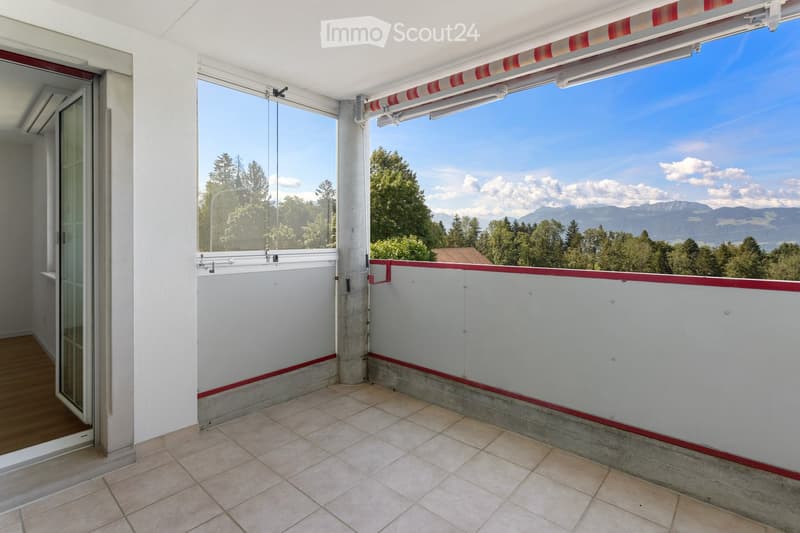 Balcony with tiled floor, white walls, red trim, large windows offering a scenic mountain view.