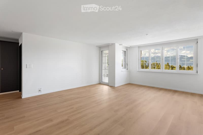 Empty room with light wooden flooring, white walls, and large windows offering a scenic view of mountains and trees.