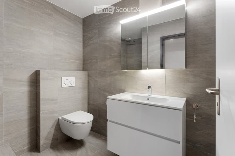 Modern bathroom with grey tiled walls, white sink with built-in cabinet, toilet, large mirror with integrated lighting, and a sleek faucet.
