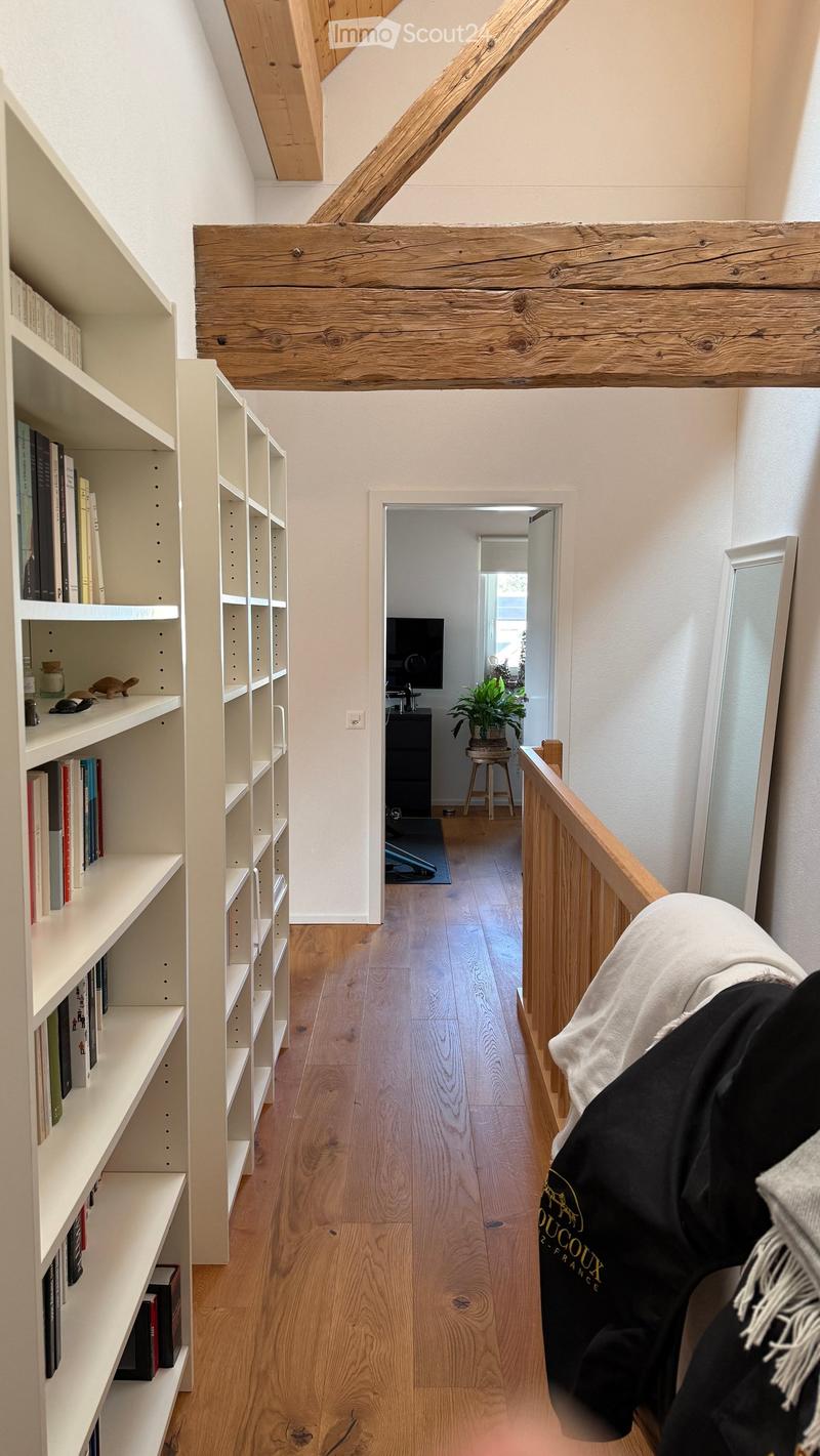 A narrow hallway with wooden floors, white walls, and exposed wooden beams. A built-in white shelving unit with books and decorative items is on the left, and a staircase leads to another room with a TV and a small plant visible in the background.