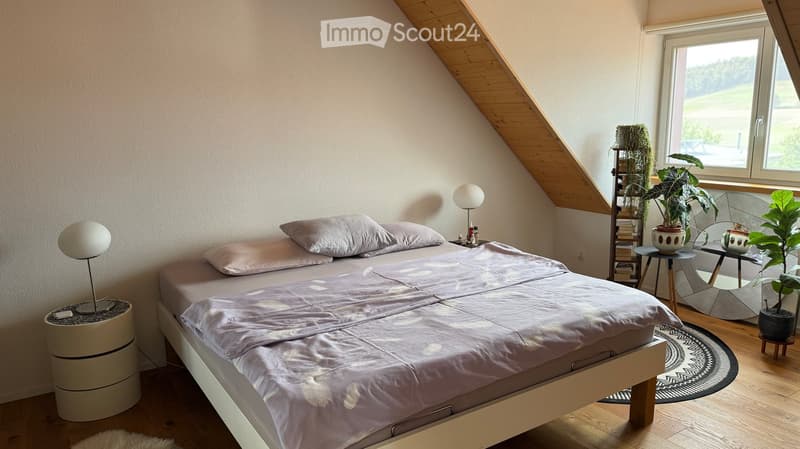 Cozy attic bedroom with wooden floors, slanted ceiling, large window, bed with light-colored bedding, bedside tables with lamps, and decorative plants.