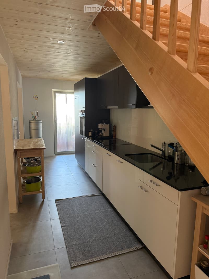 Modern kitchen with white cabinets, black countertops, and stainless steel appliances. Features a sink, coffee maker, and a small wooden stool with pots.