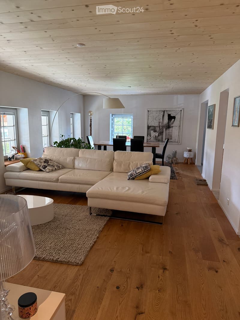 Modern living room with light-colored wooden flooring, a large white sectional sofa, a coffee table, a floor lamp, and a dining area with chairs and a table.