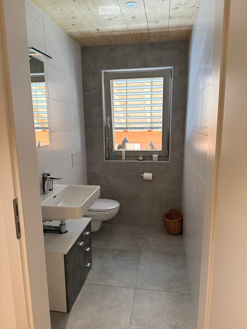 Modern bathroom with grey tiles, white sink, toilet, mirror, and wicker basket on the floor.