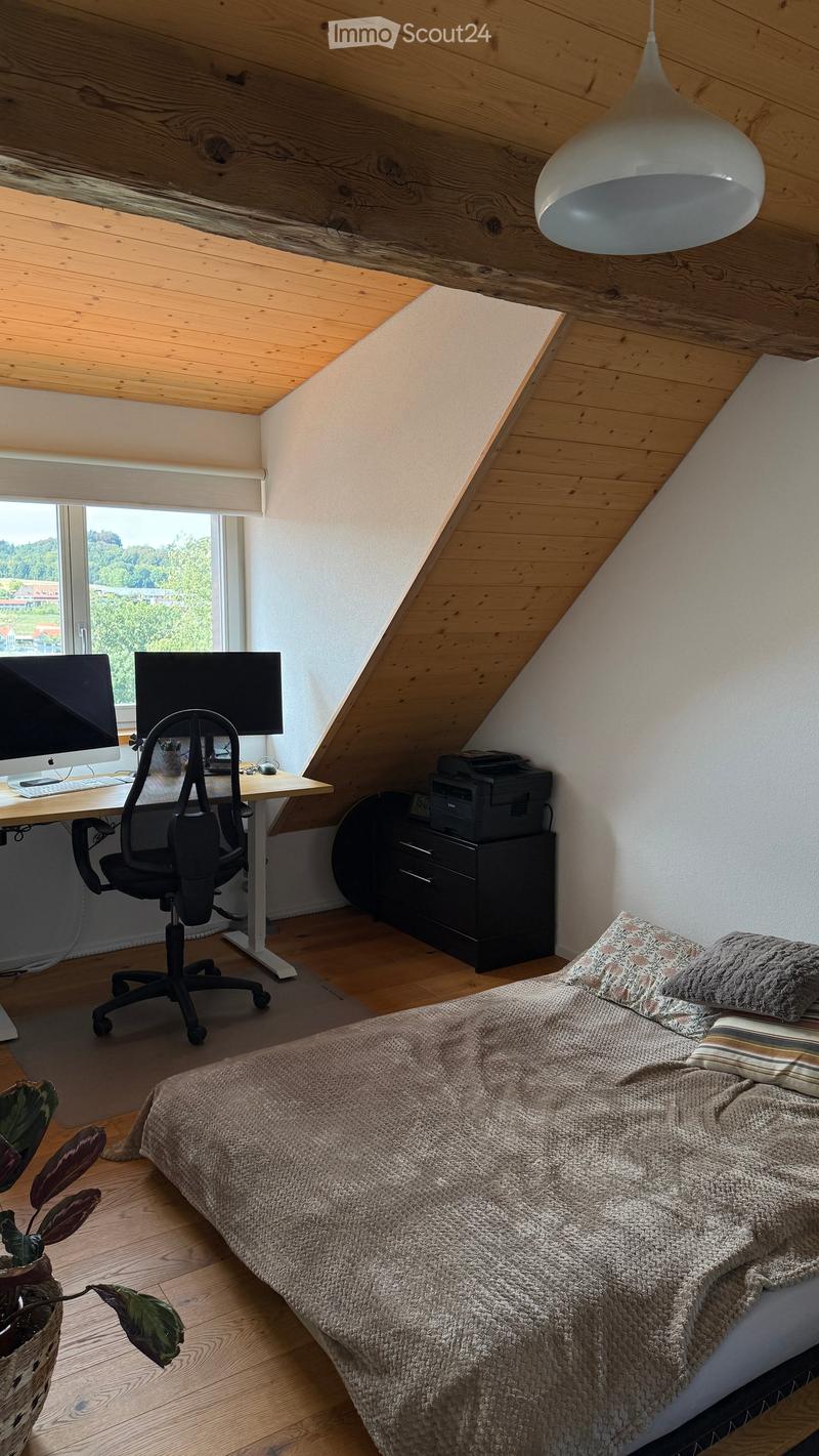 Cozy attic bedroom with a slanted wooden ceiling, large window, bed with textured bedding, desk with computer and chair, printer on a cabinet, and a potted plant near the bed.