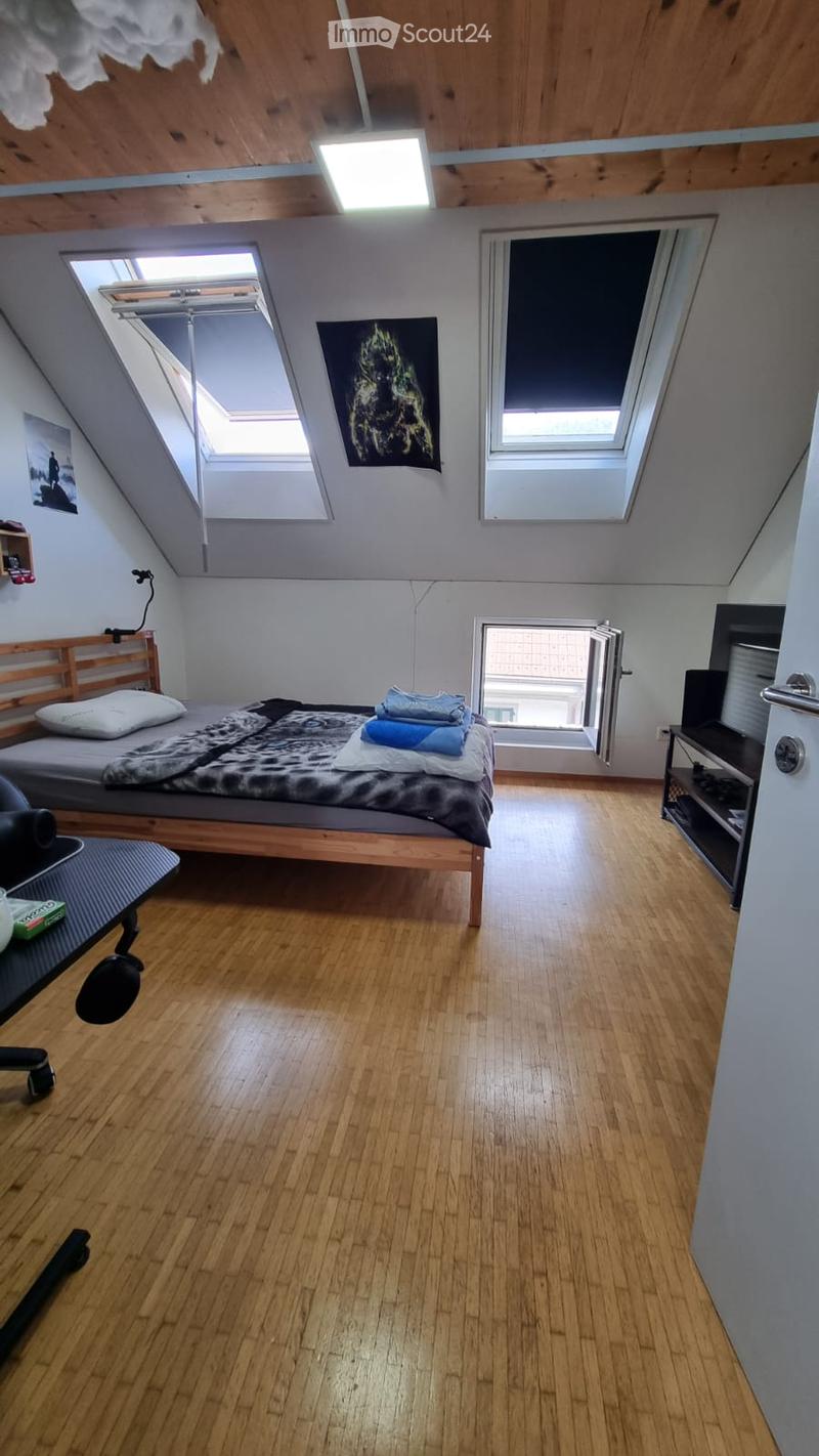 Cozy attic bedroom with wooden floors, slanted wooden ceiling, skylights, bed with pillows and blankets, chair, small table, TV screen, and artwork on walls.