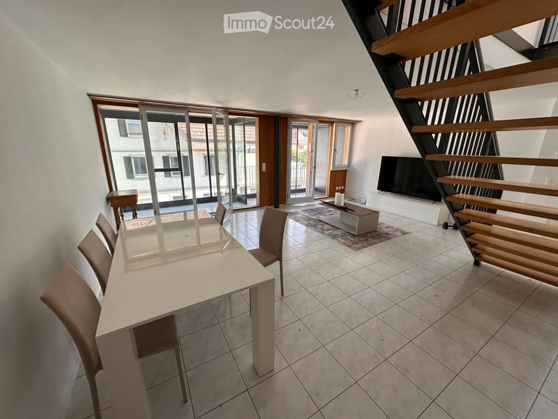 Bright, modern dining area with a white dining table, four chairs, a television on a stand, and a small wooden table near the sliding glass doors leading to a balcony.
