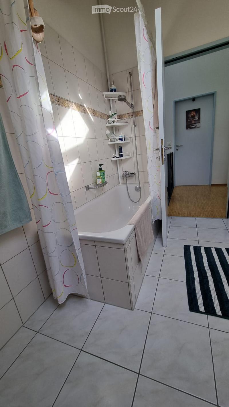 Bathroom with a bathtub, shower curtain, towel, and bathroom shelf with bottles. The floor is tiled, and there is a black and white striped rug near the door.