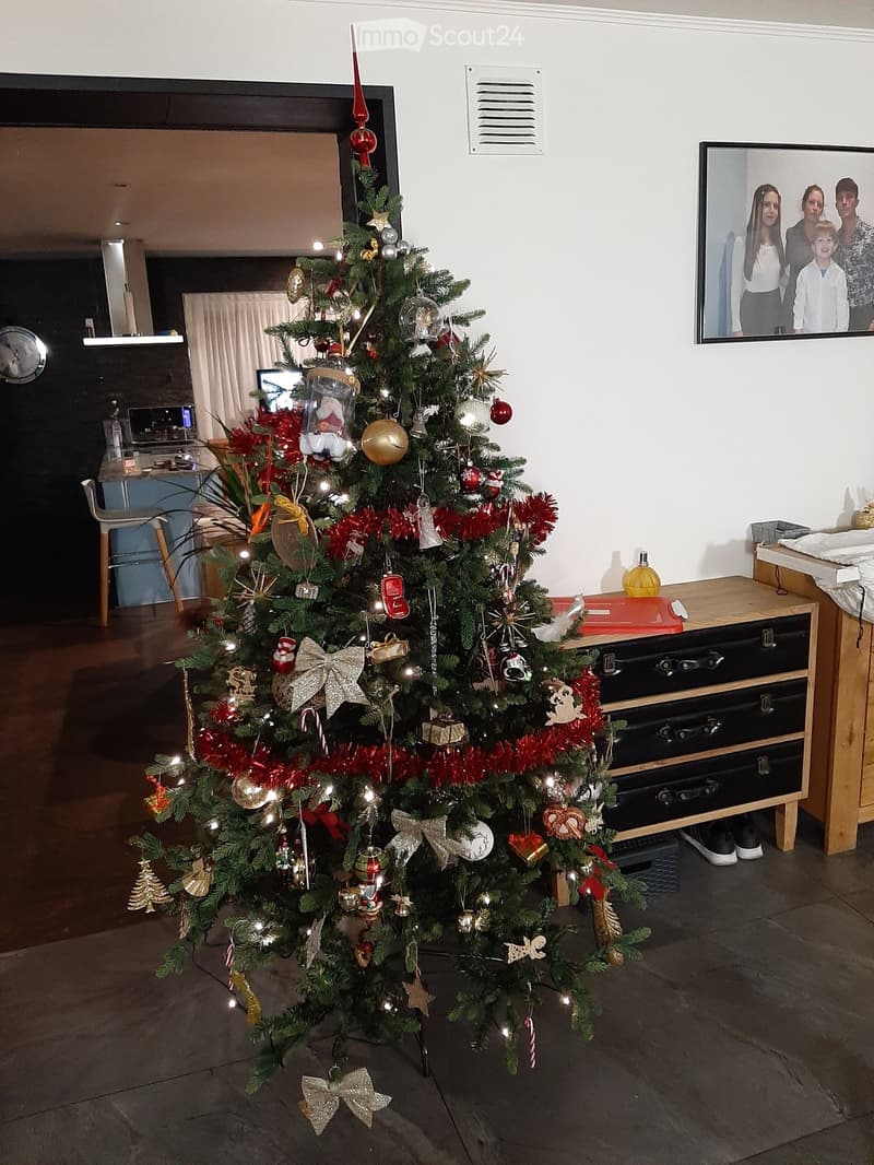 A large, decorated Christmas tree stands in the center of a room, surrounded by a chair, a small table, and a screen displaying a family photo. The tree is adorned with colorful ornaments, lights, and tinsel.