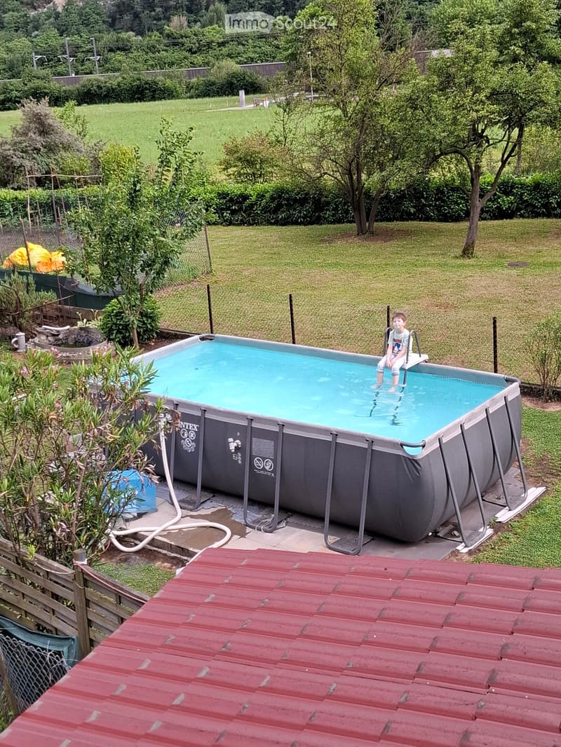 Backyard with a rectangular swimming pool, surrounded by grass, trees, and a wooden fence. A child is sitting on the edge of the pool.