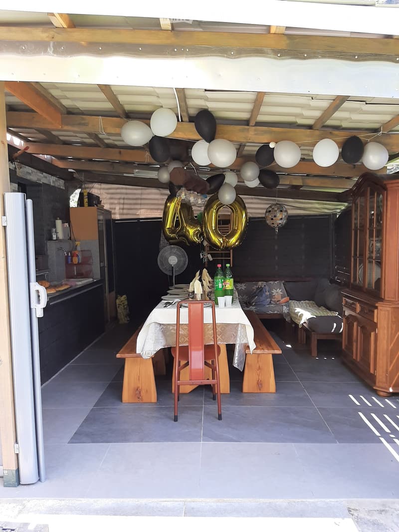 A covered outdoor dining area with a table, chairs, benches, a sofa, and a wooden cupboard. Balloons and a fan are visible, and the area is decorated for a celebration.