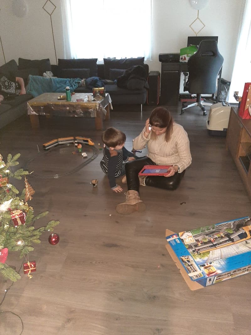 A cozy living room with a sofa, coffee table, and a laptop on a table. A woman is sitting on the floor with a child, and there is a Christmas tree with ornaments nearby.