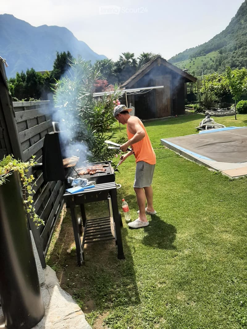 A man is grilling sausages on a barbecue in a grassy yard with a wooden shed and mountains in the background.