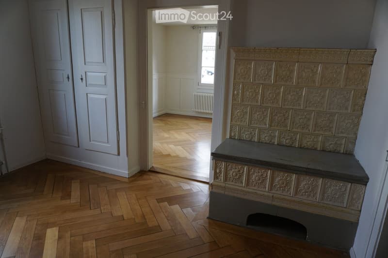 Empty hallway with wooden flooring, white walls, and a decorative fireplace with ornate tiles. A door leads to another room with a radiator and window.