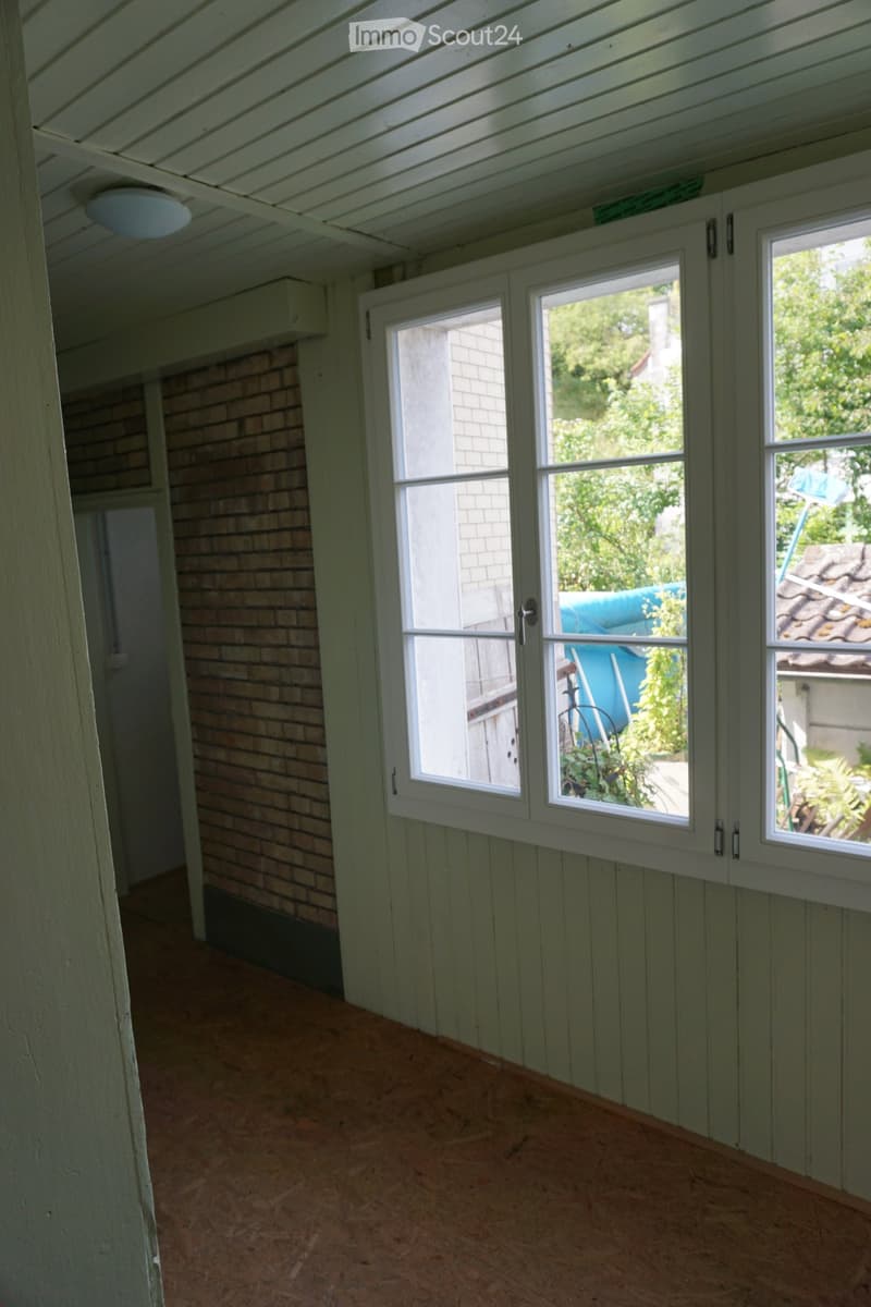 Empty room with wooden flooring, white paneled walls, and large windows offering a view of the garden and neighboring house.