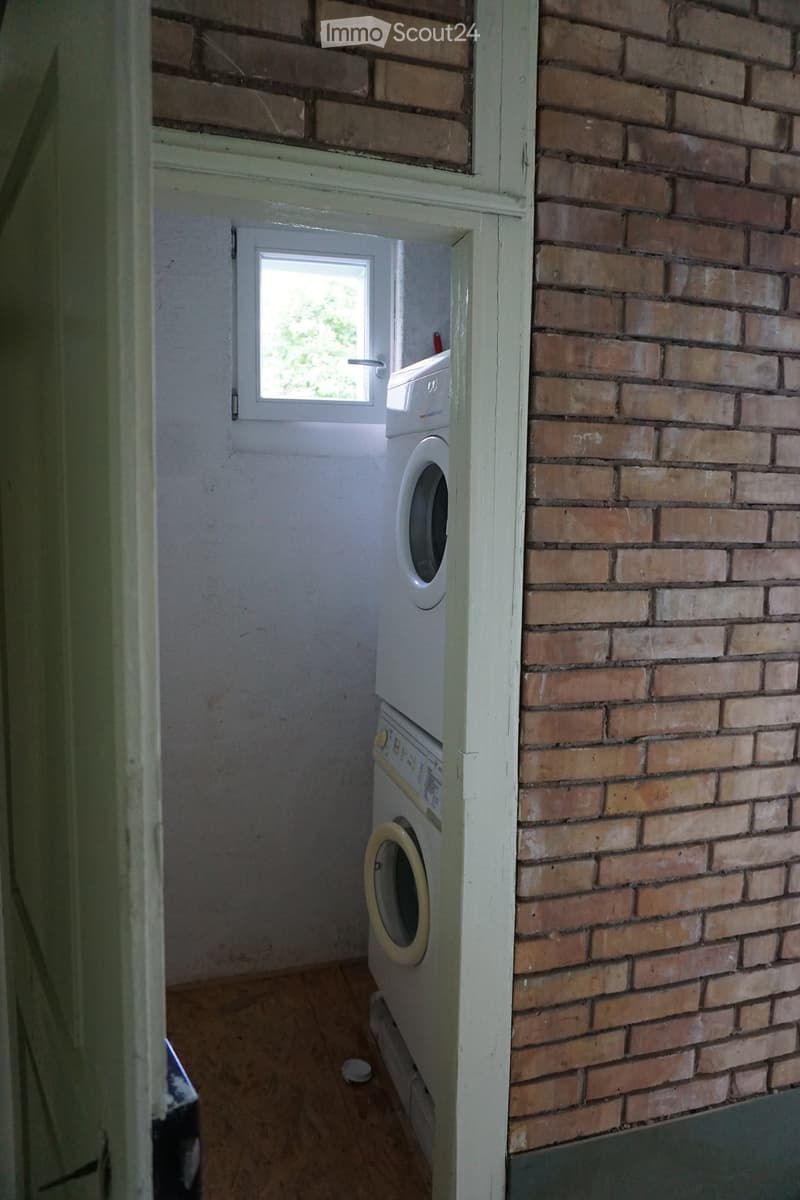 A small room with a washing machine, brick wall, wooden floor, and a window with white frame.