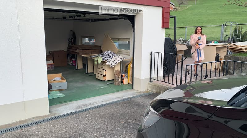 A car park with garage, outdoor tables, and a woman standing in front of it