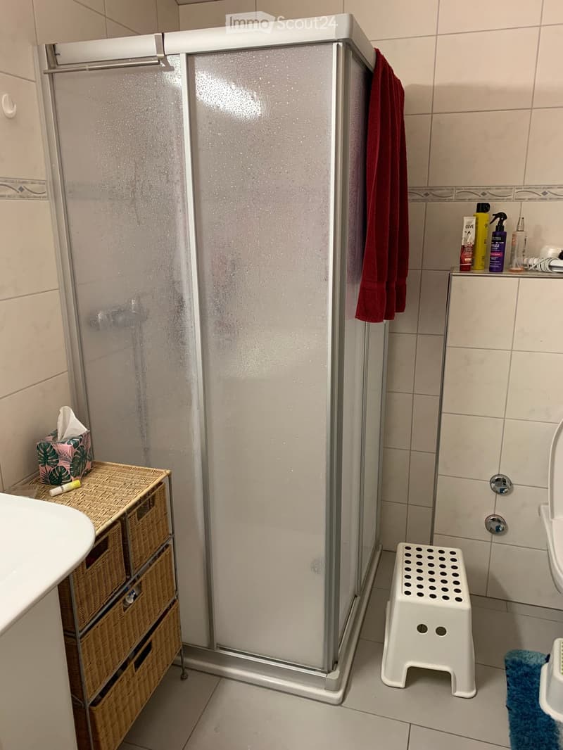 A bathroom with white tiles, a white sink, a shower stall, a red towel hanging on a rack, a wicker basket on the side of the sink, a toilet, a soap dispenser, a small stool in front of the toilet, a tissue box on top of the basket, a white toilet paper ho