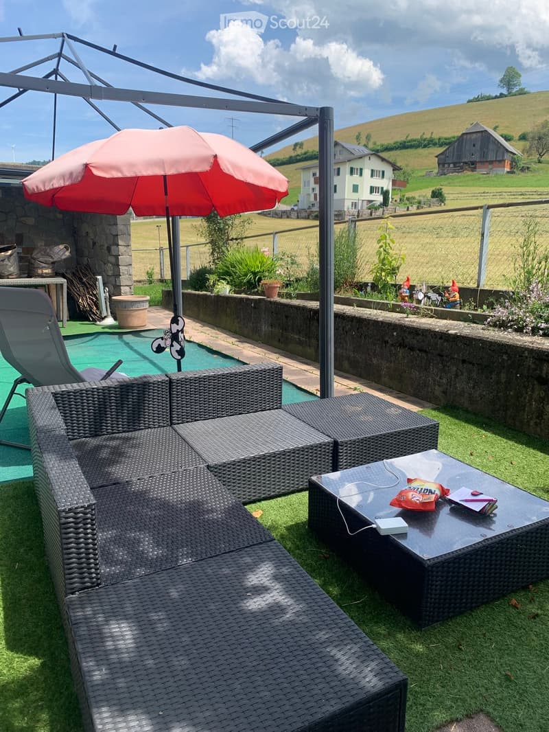 A rattan sofa, a coffee table, and an umbrella are placed on a well-maintained lawn with stone walls in the background. Decorative figurines and plants add to the ambiance. In the distance, a field and a white house are visible.