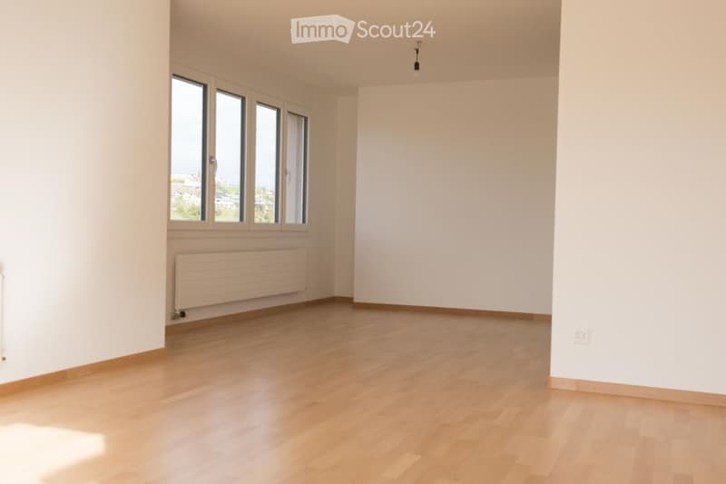 Empty room with light wooden flooring, white walls, large windows, and a radiator. No furniture or equipment present.