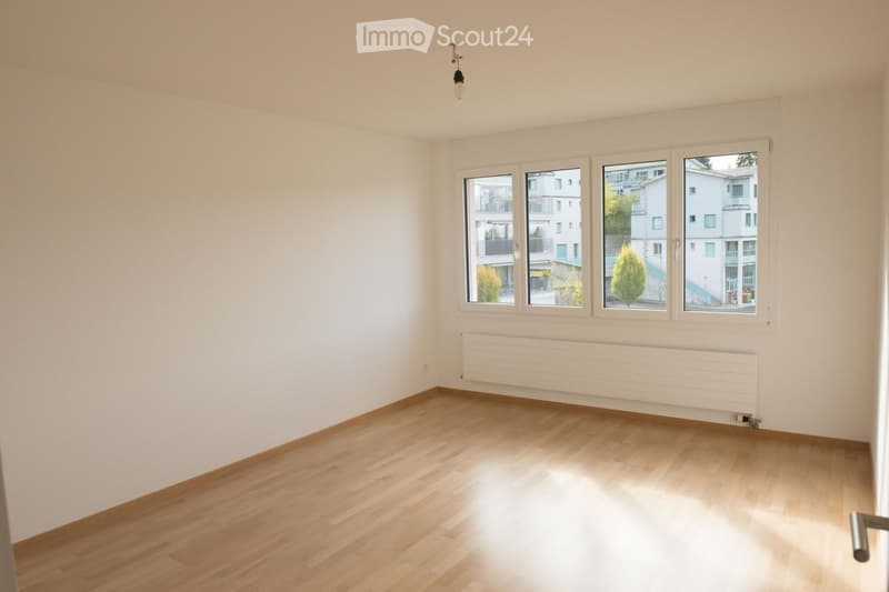 Empty room with light wooden flooring, white walls, large windows, and a radiator below the windows.